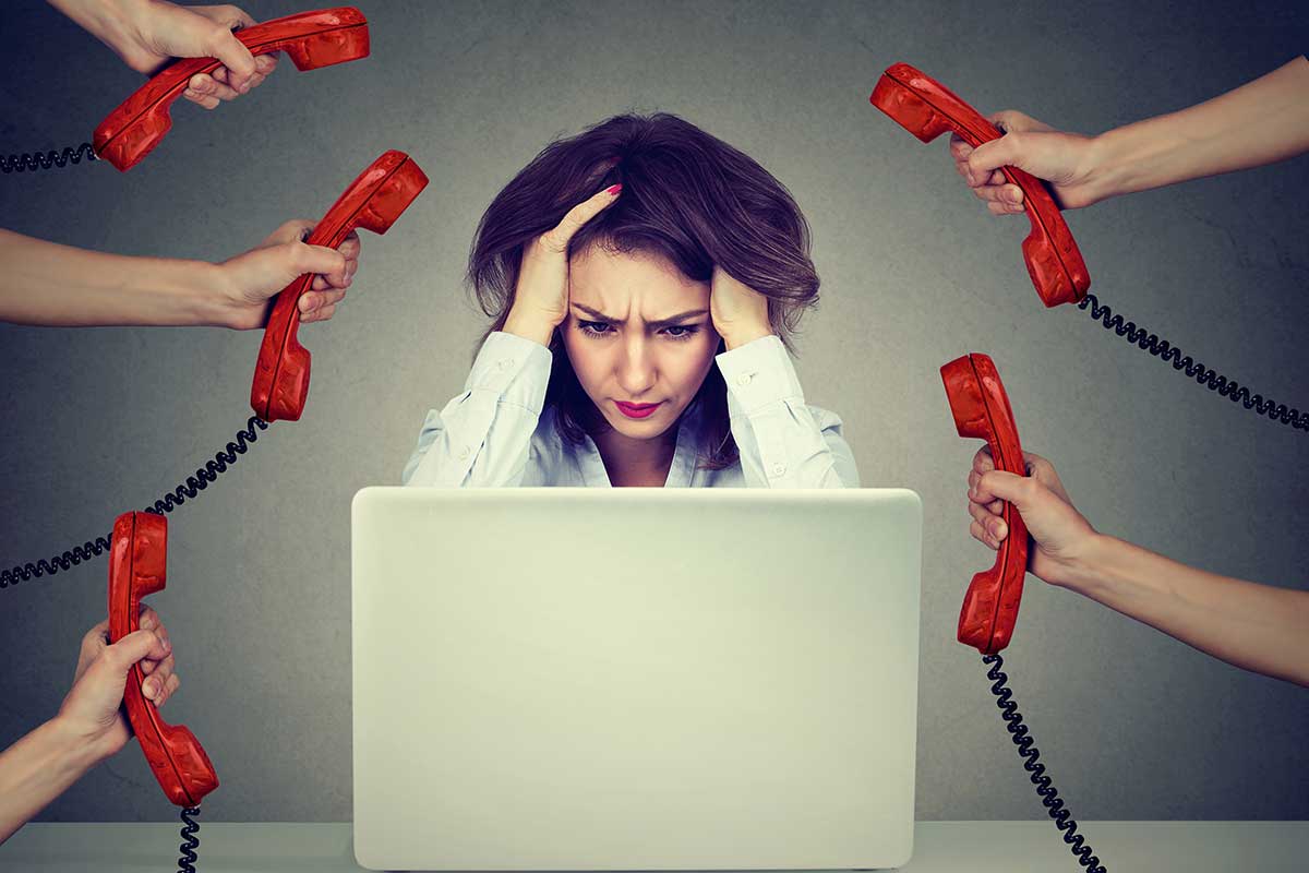 stressed out business woman with hands on head, looking at a laptop screen, and 5 hands holding phone handsets to her