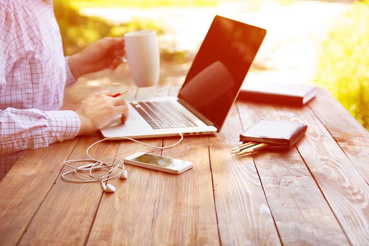 business person holding a coffee mug and working remote from outdoor table from laptop and mobile phone